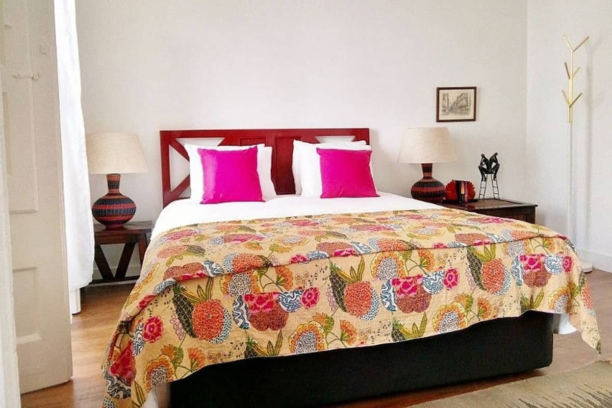 Un lit avec une couvre-lit à motifs floraux, deux oreillers blancs et deux oreillers roses. Il y a deux tables de chevet avec des lampes, et un petit objet décoratif sur l’une d’elles.