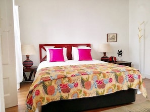 Un lit avec une couvre-lit à motifs floraux, deux oreillers blancs et deux oreillers roses. Il y a deux tables de chevet avec des lampes, et un petit objet décoratif sur l’une d’elles.