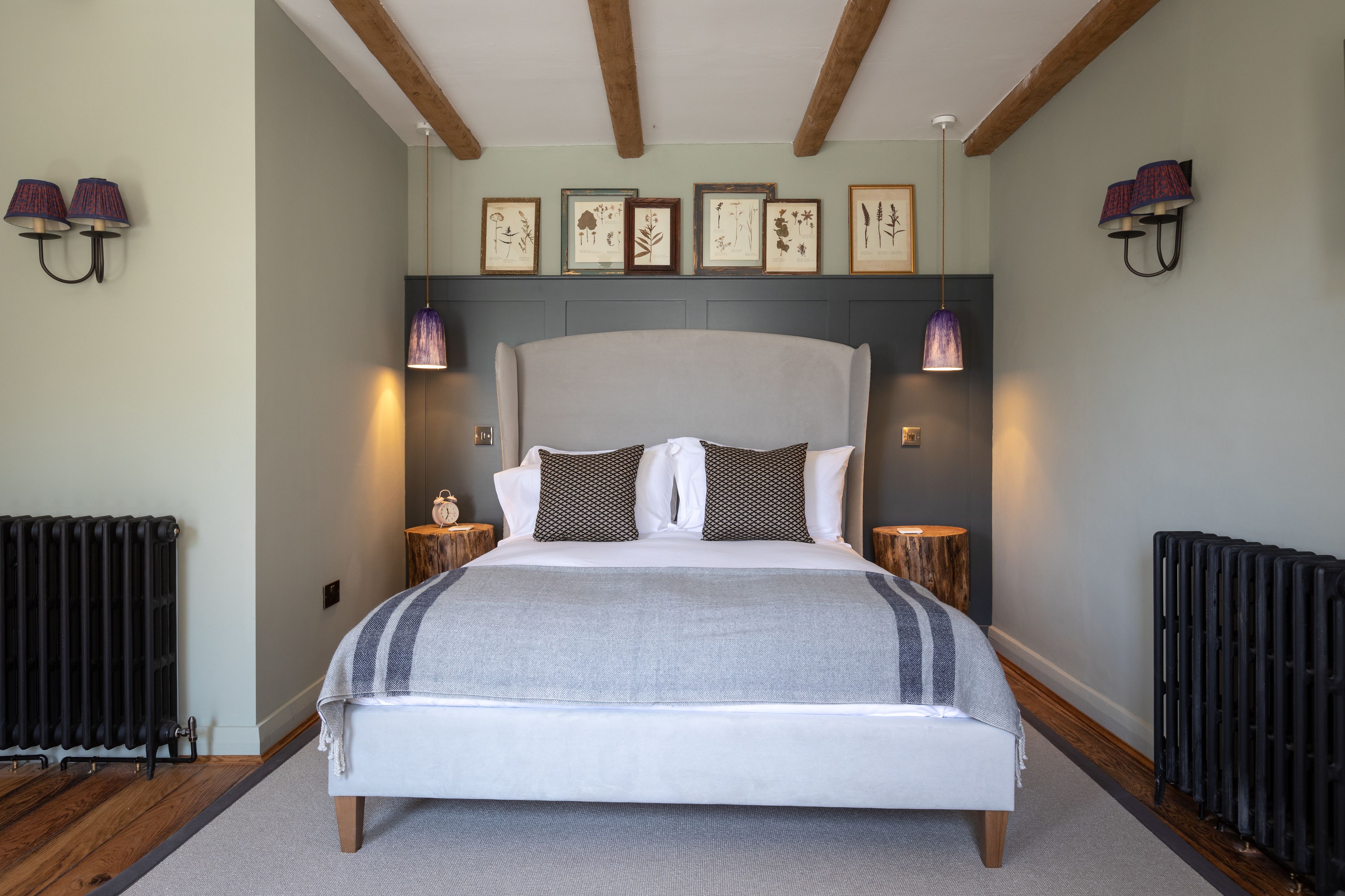 A bedroom with a large bed, wooden beams, framed botanical prints, and wall-mounted lamps.