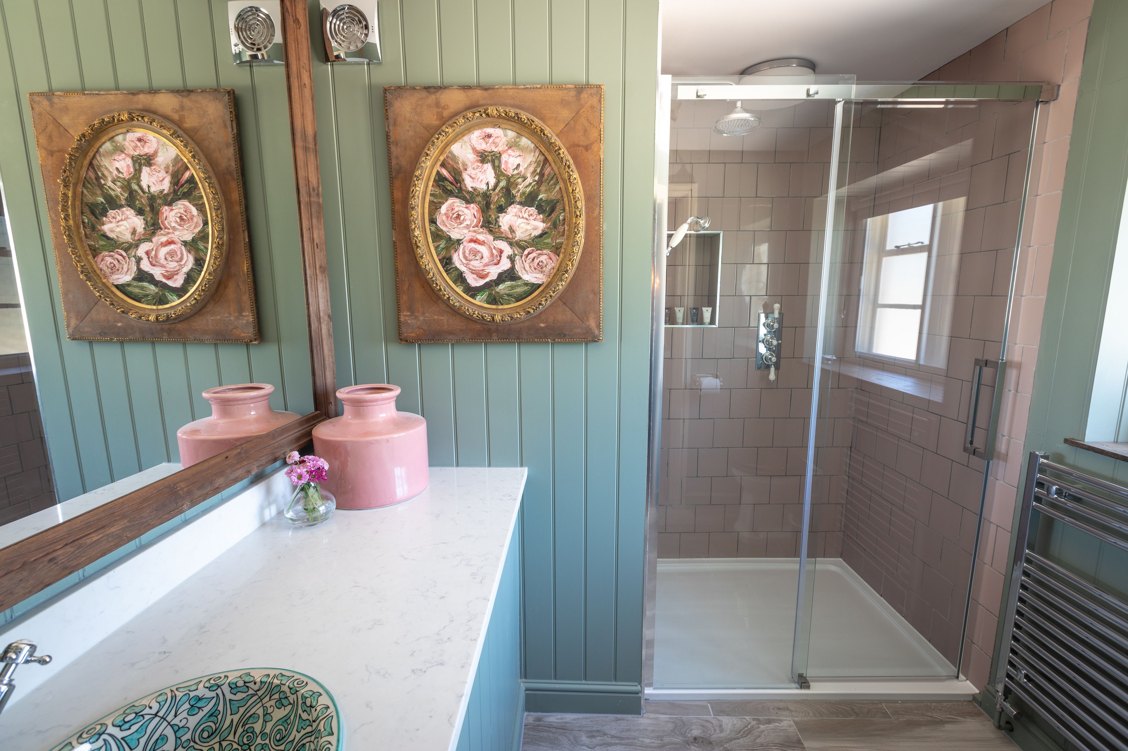 Une salle de bain avec une douche à parois de verre, un meuble-lavabo avec un miroir et deux tableaux à motifs floraux encadrés.