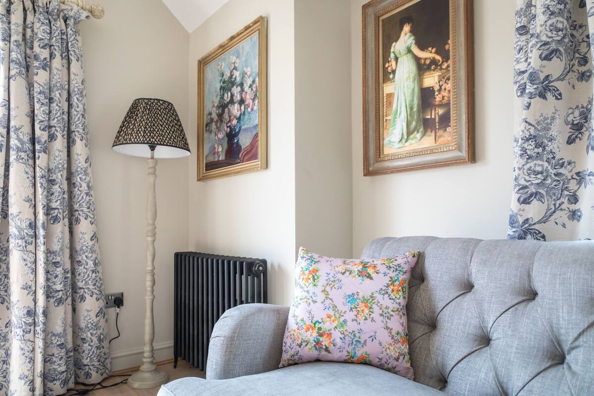 A living room with a grey sofa, floral patterned curtains, a standing lamp, and two framed paintings on the wall.