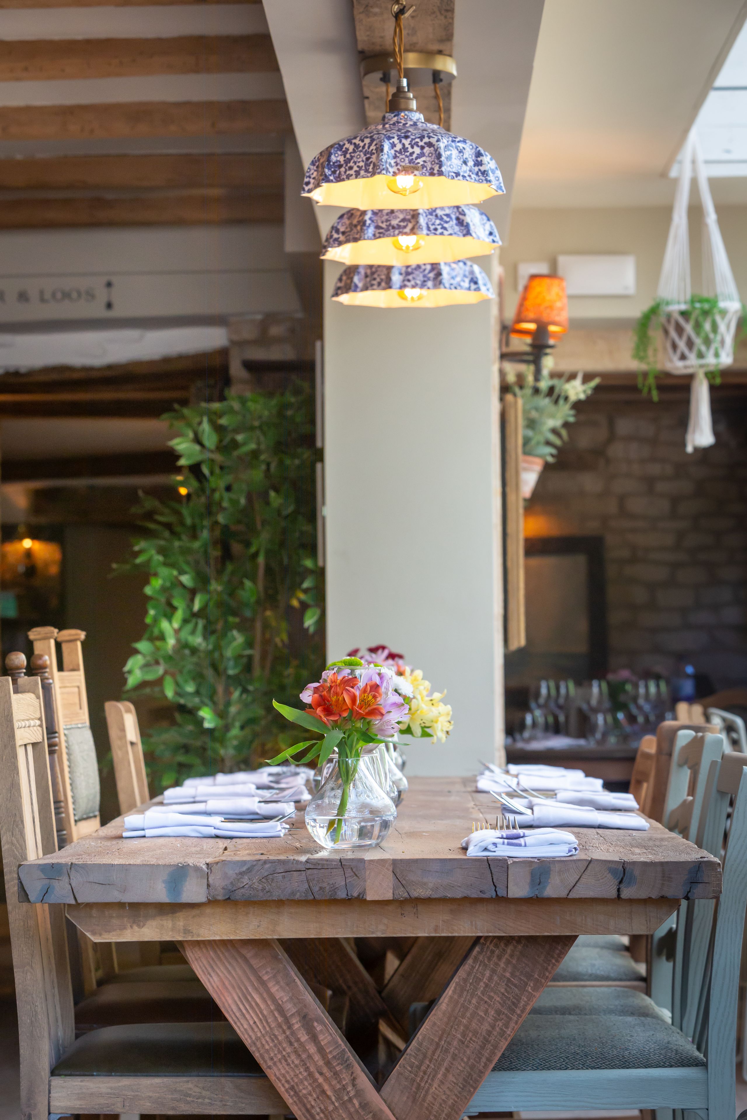 Una mesa de comedor de madera con un florero, dispuesta con servilletas y cubiertos blancos, bajo una lámpara colgante de diseño azul y blanco.