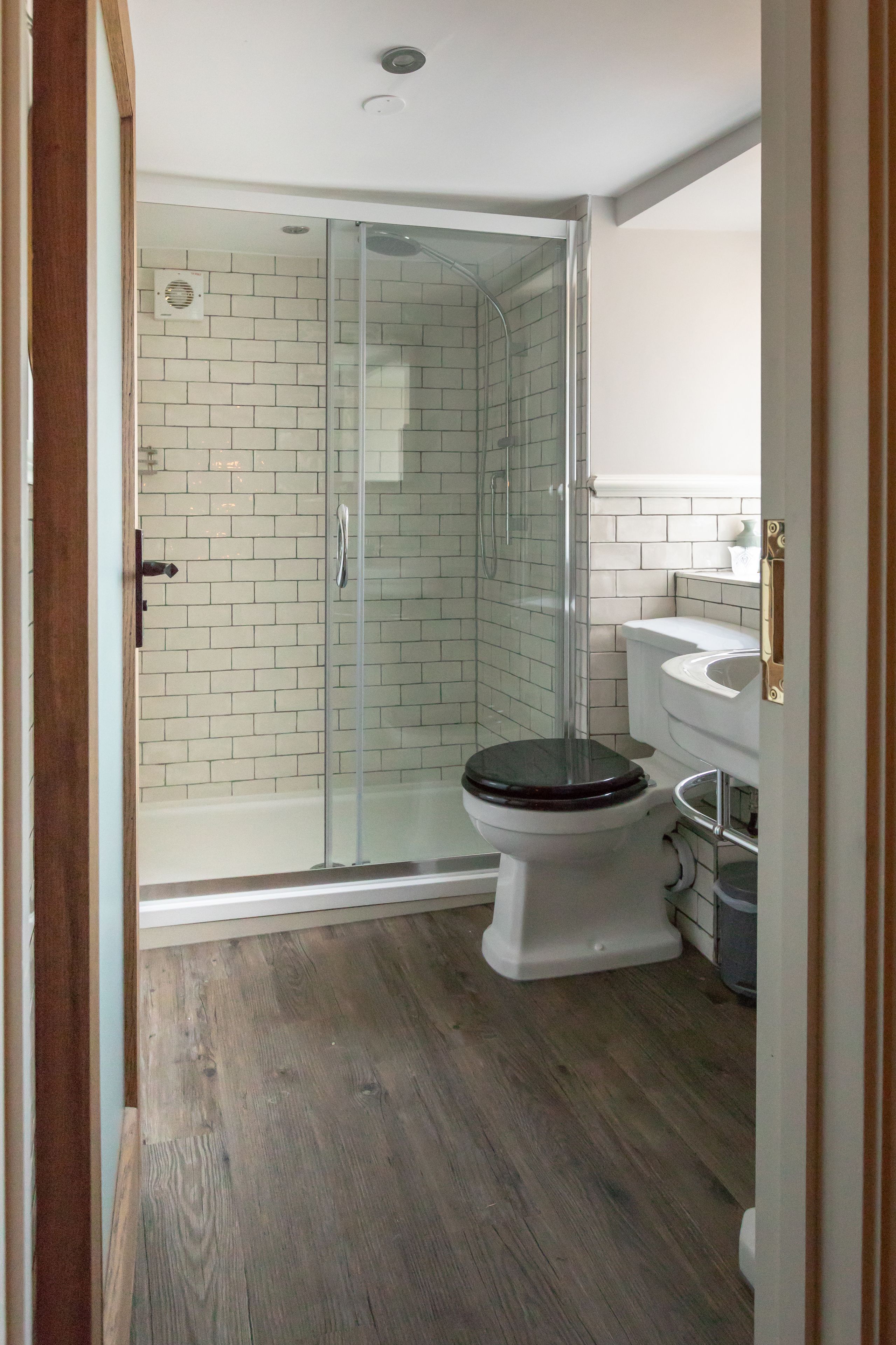 A bathroom with a glass-enclosed shower, white tiled walls, a wooden floor, and a white toilet with a black seat.