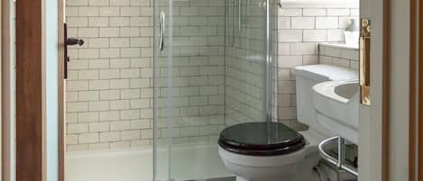 A bathroom with a glass-enclosed shower, white tiled walls, a wooden floor, and a white toilet with a black seat.