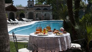 A poolside dining area with a table set for a meal, surrounded by lounge chairs and a stone building in the background. - La Villa (Giardini Naxos)
