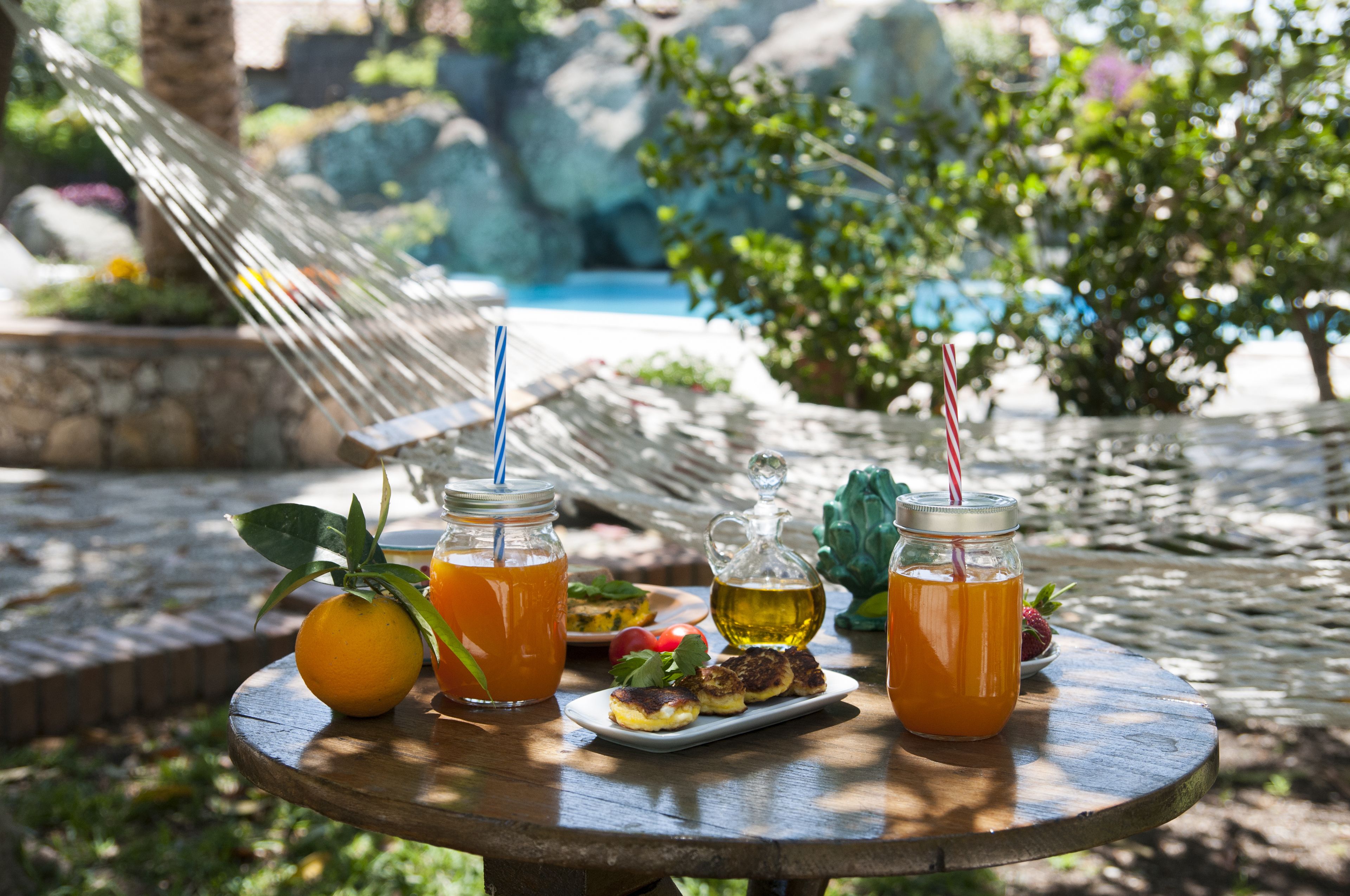 A wooden table with drinks, a fruit, and a small dish of food in a garden setting.