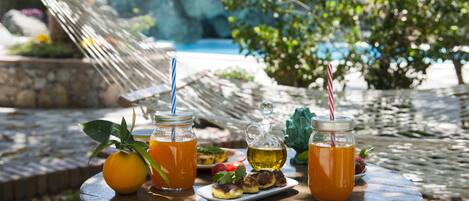 A wooden table with drinks, a fruit, and a small dish of food in a garden setting.