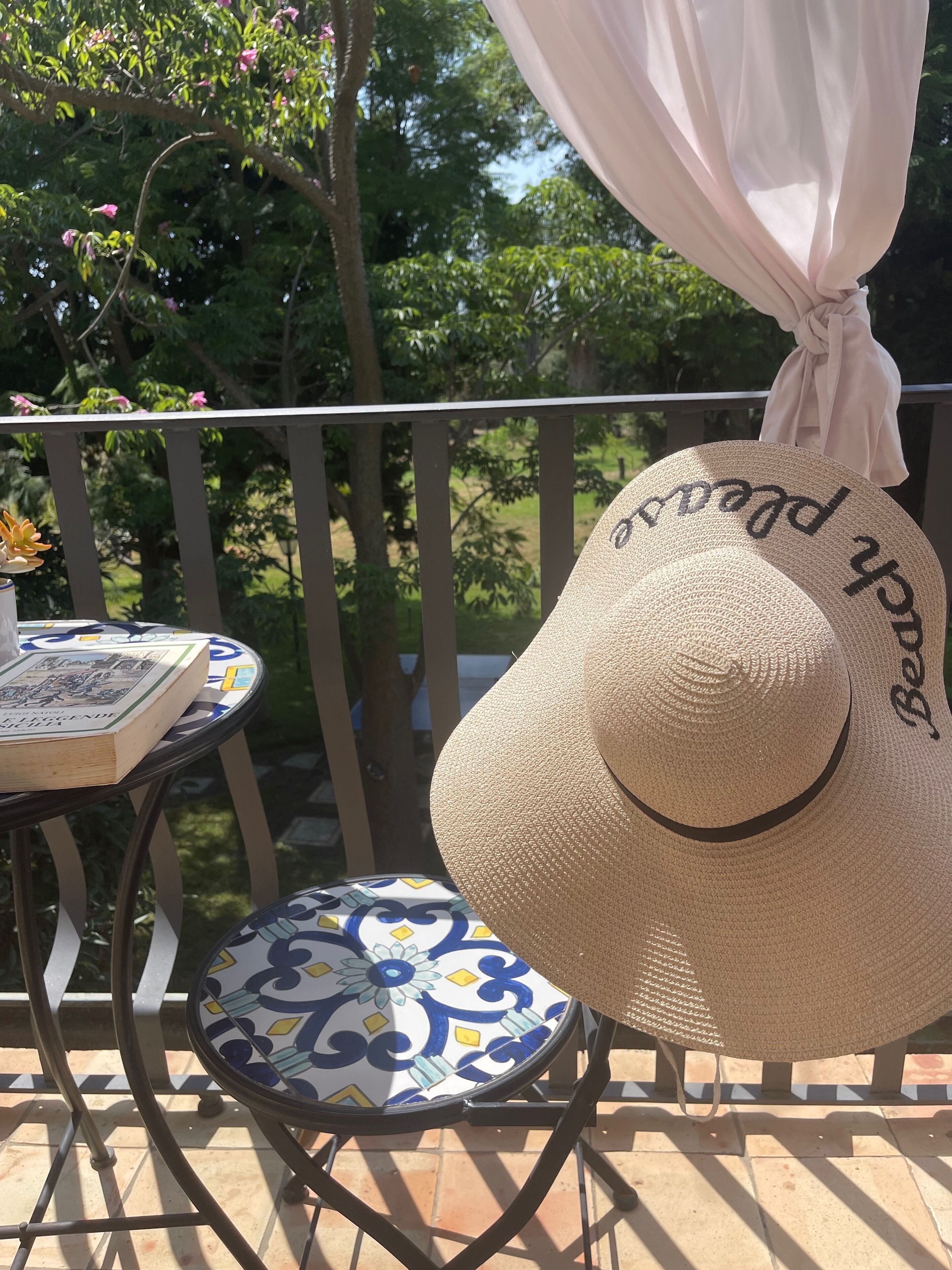 Un balcon agrémenté d’un grand chapeau de paille, une table avec un livre et une nappe à motifs.