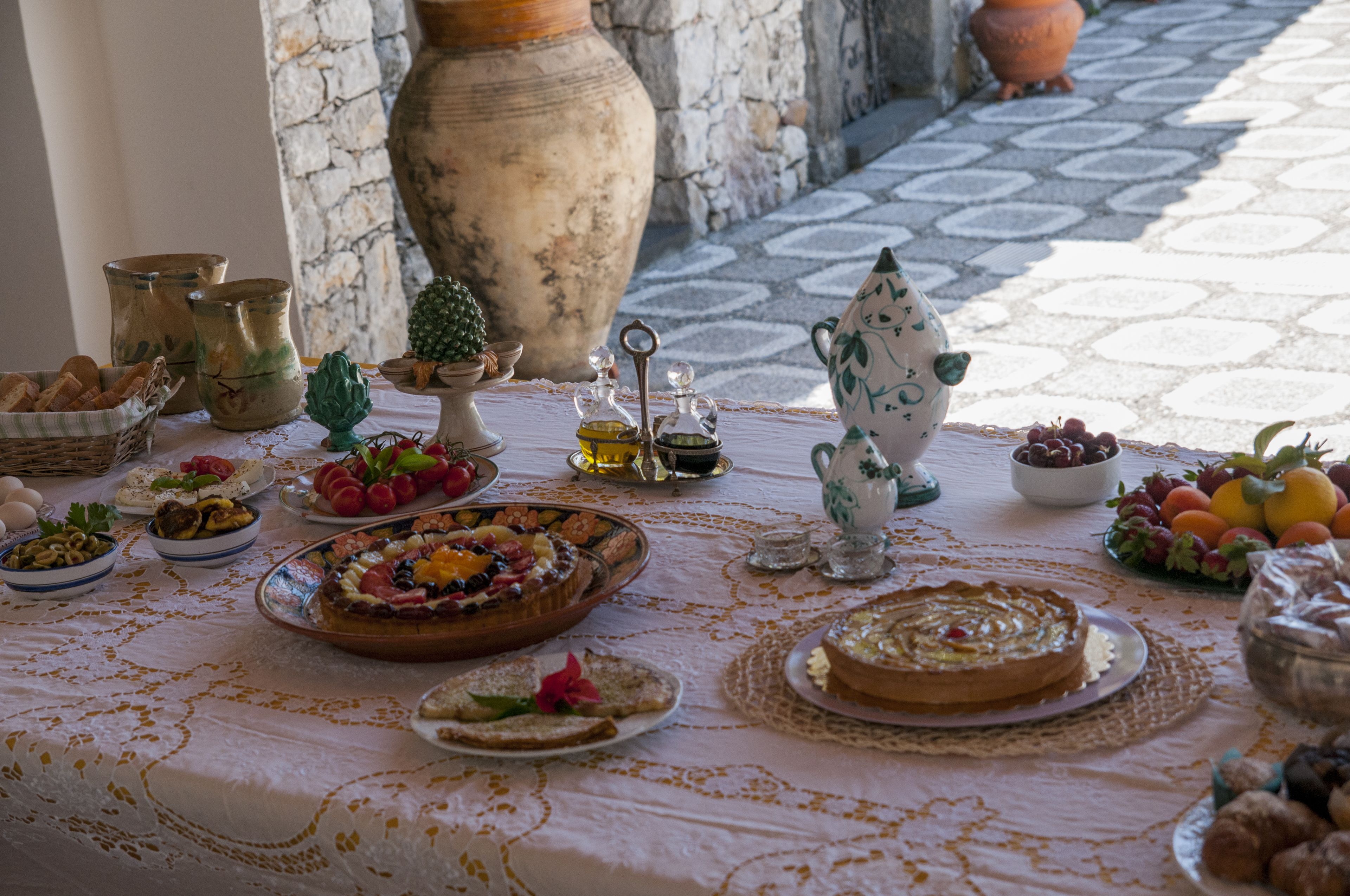 Une table dressée avec divers plats, notamment un assortiment de fruits, du pain et un gâteau, dans une pièce aux murs de pierre et au sol carrelé.