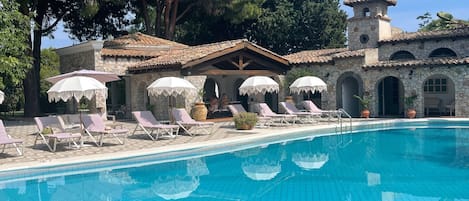 Una piscina con tumbonas y sombrillas, rodeada de edificios de piedra y árboles.