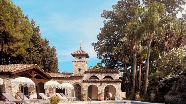 A pool area with a stone building, lounge chairs, and a person reading a book. - La Villa (Giardini Naxos)