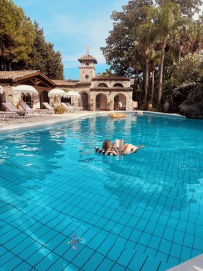 Zona de piscina con un edificio de piedra, sillas de descanso y una persona leyendo un libro.
