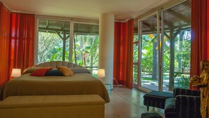 A spacious bedroom with a large bed, a seating area, and a view of lush greenery through large windows. - La Villa (Giardini Naxos)