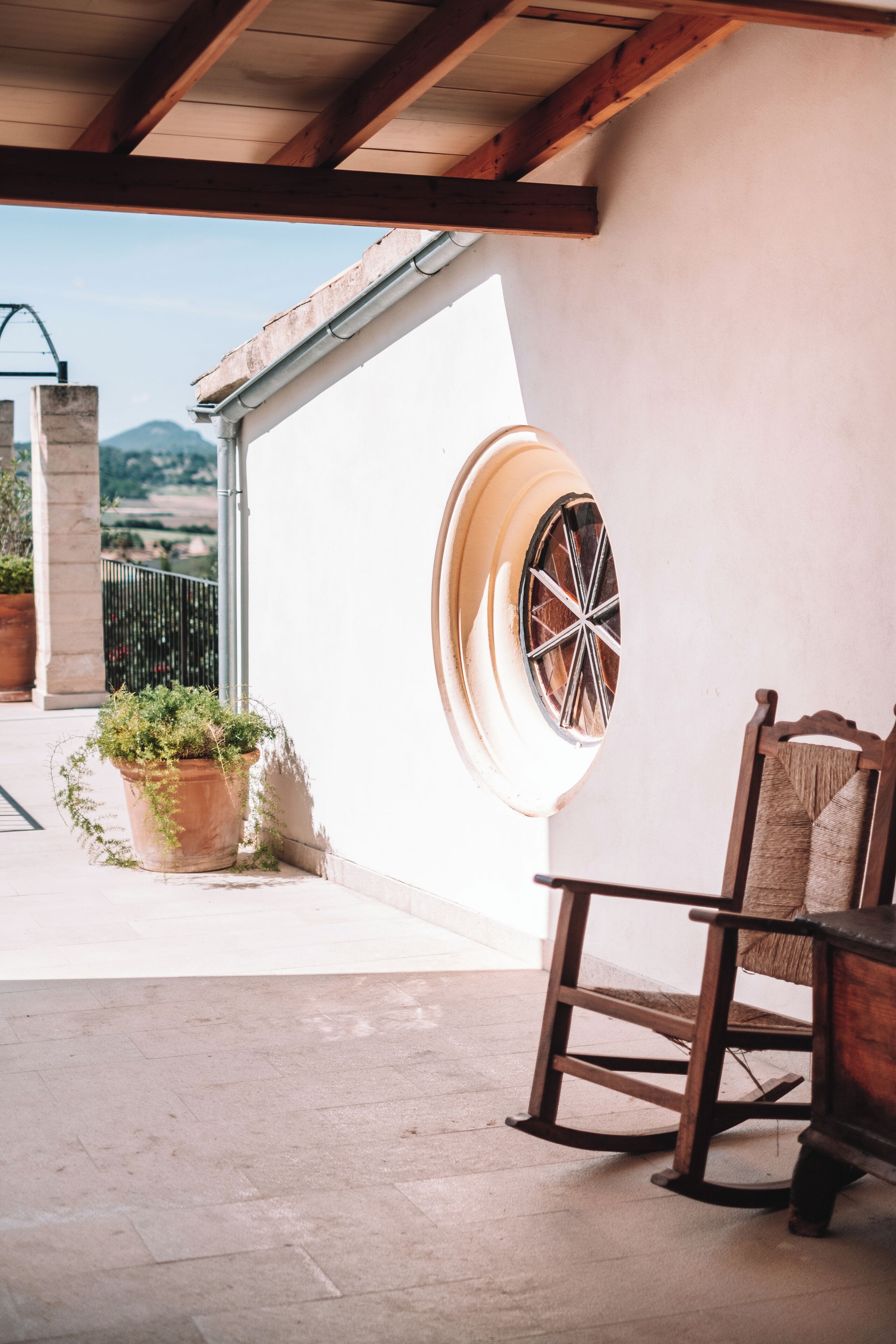 A covered patio area with a wooden rocking chair, a potted plant, and a large window with a decorative grille.