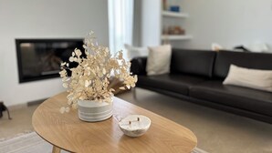 Un saló modern amb una taula baixa de fusta, un vas de flors seques i blanc, i un bol blanc. Es veu un sofà de tons foscos i una llar de foc al fons.