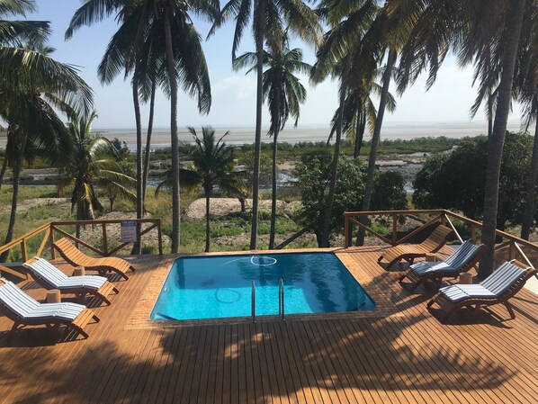 Une terrasse en bois avec une piscine, des chaises longues et une vue sur la plage et les palmiers.