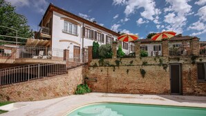 A pool area with a brick wall, a white building with a balcony, and red and white umbrellas. - Il Pozzo Fiorito Residence (Cossombrato)