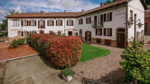 A white building with brown shutters and a red-tiled roof, surrounded by a brick courtyard and green lawn. - Il Pozzo Fiorito Residence (Cossombrato)