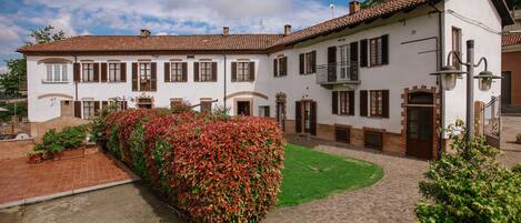A white building with brown shutters and a red-tiled roof, surrounded by a brick courtyard and green lawn.