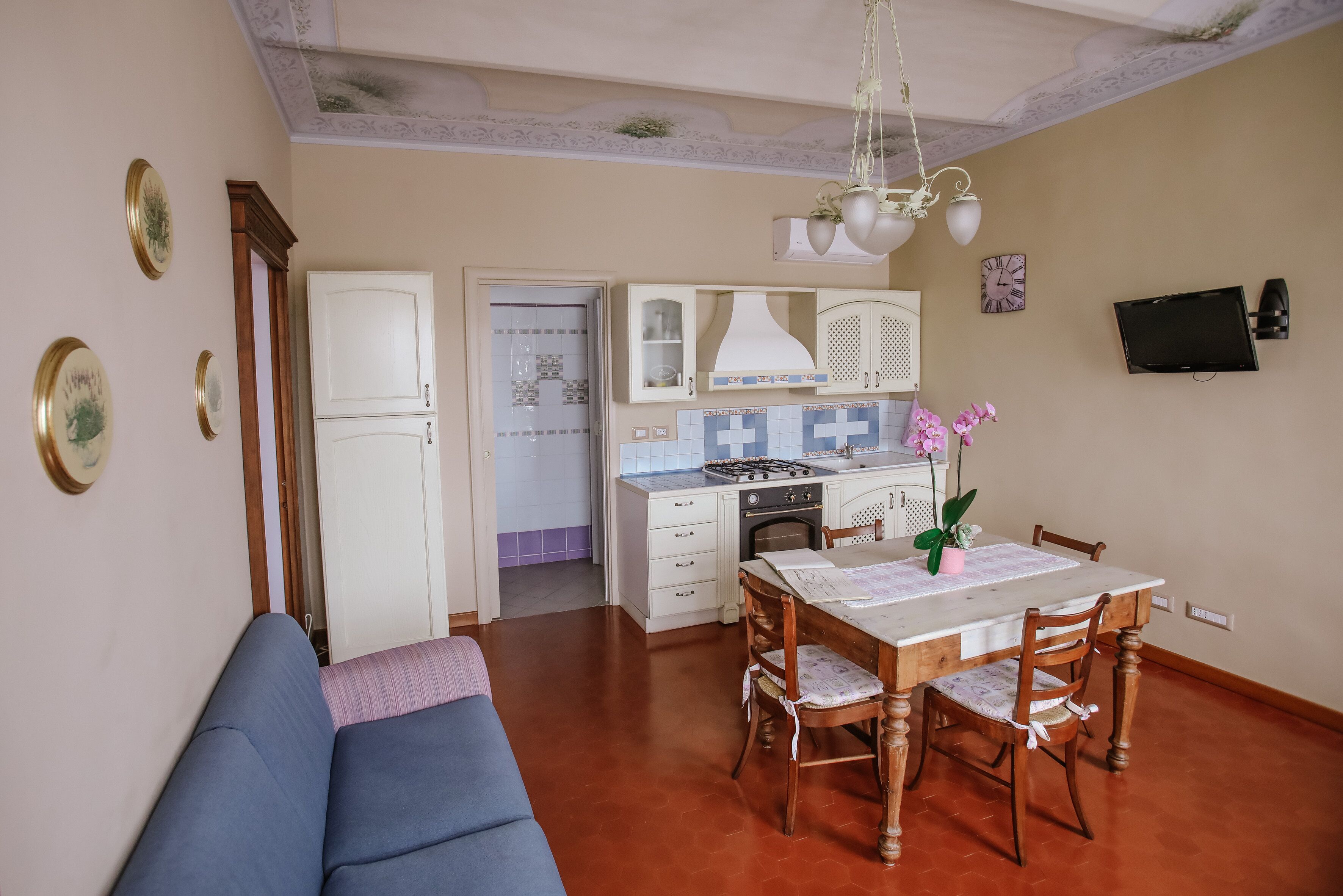 A dining area with a wooden table and chairs, a sofa, a kitchen with white cabinets, and a TV mounted on the wall.