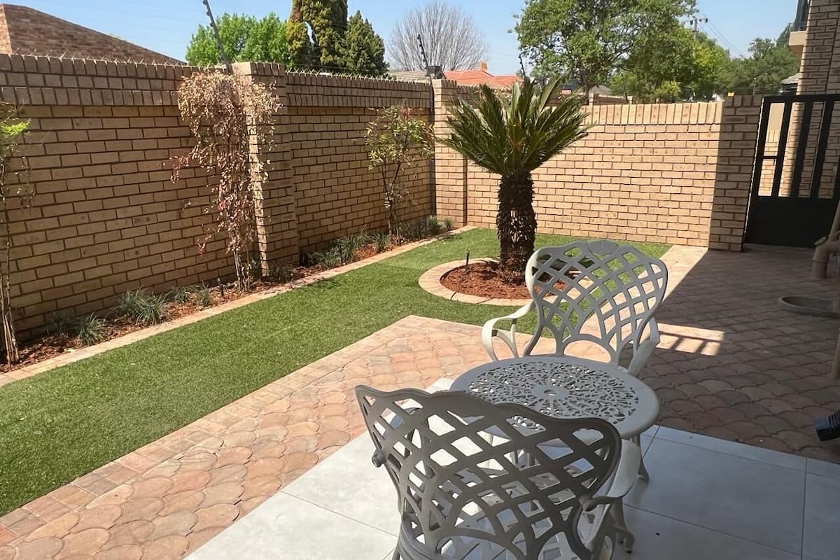 A patio area with two white metal chairs and a small round table, surrounded by a brick wall and a well-maintained lawn.