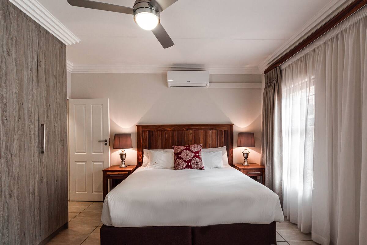 A hotel room with a large bed, wooden headboard, two bedside tables with lamps, a ceiling fan, and an air conditioning unit.