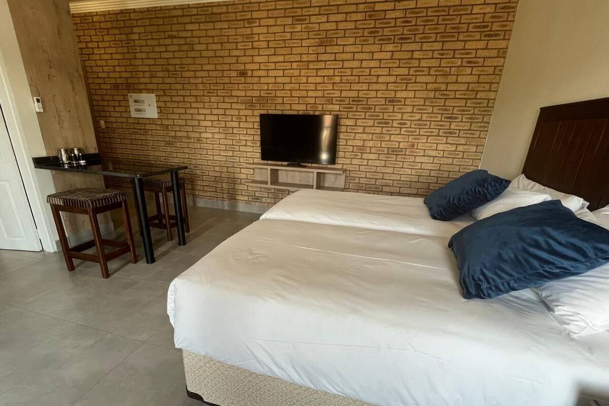 A hotel room with a large bed, a TV mounted on a brick wall, a small table, and a chair.