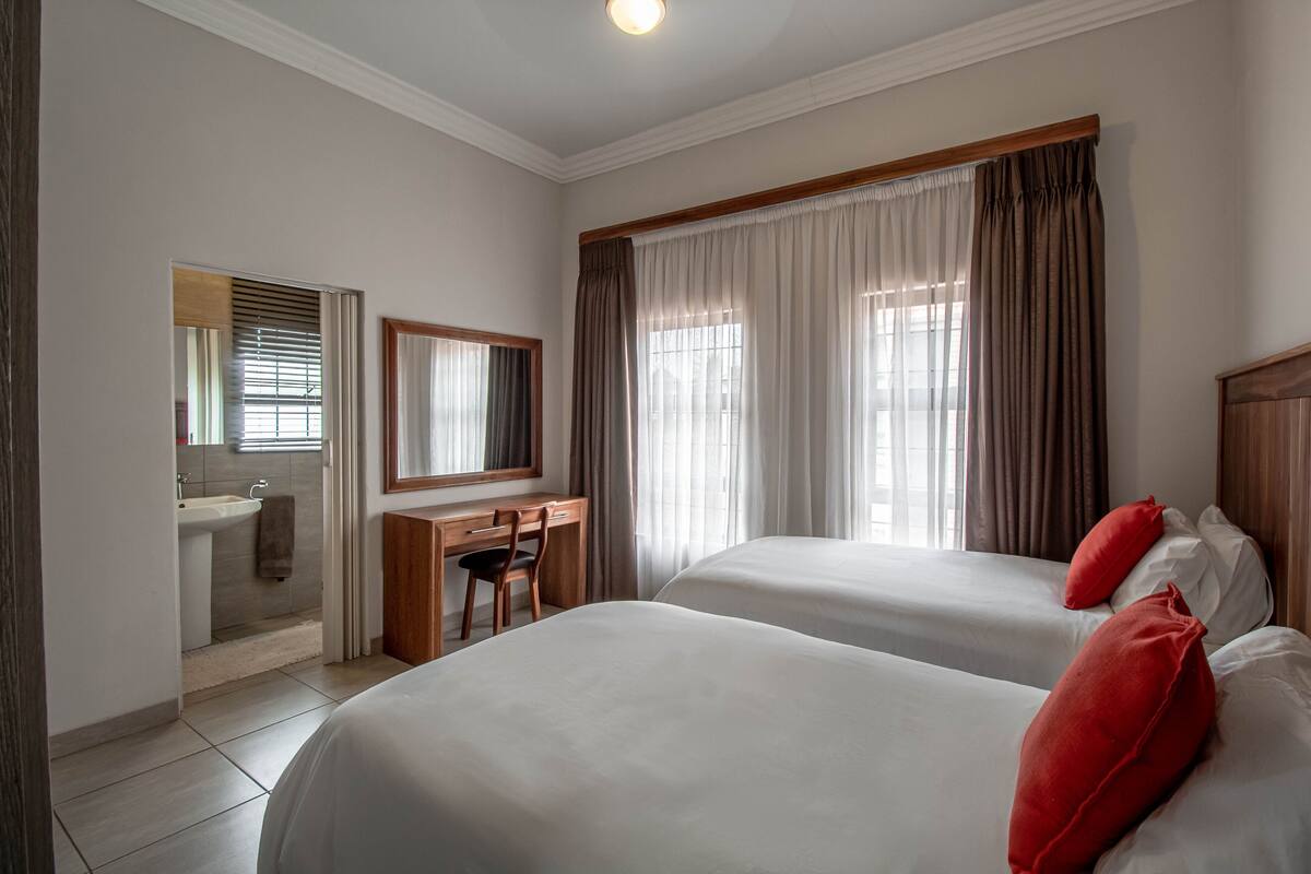 A hotel room with two beds, a desk, and a bathroom with a sink and mirror.