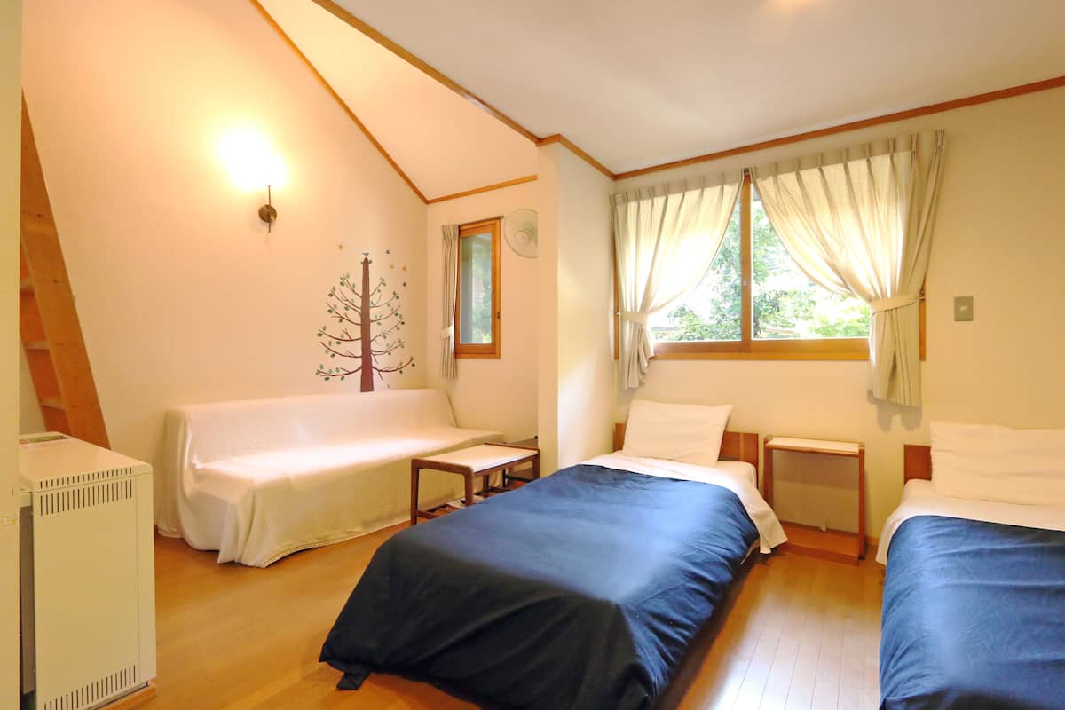 A room with two beds, a small table, a chair, a radiator, and a window with curtains.