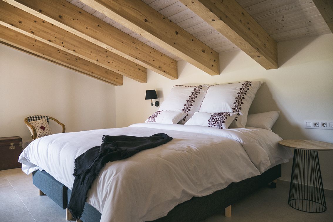 A bedroom with a bed, wooden beams, a chair, a lamp, and a small table.