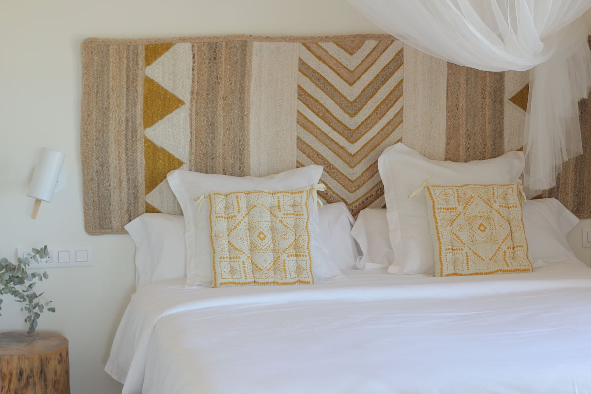 A neatly made bed with white linens and decorative pillows, a wooden nightstand with a plant, and a wall-mounted light fixture.
