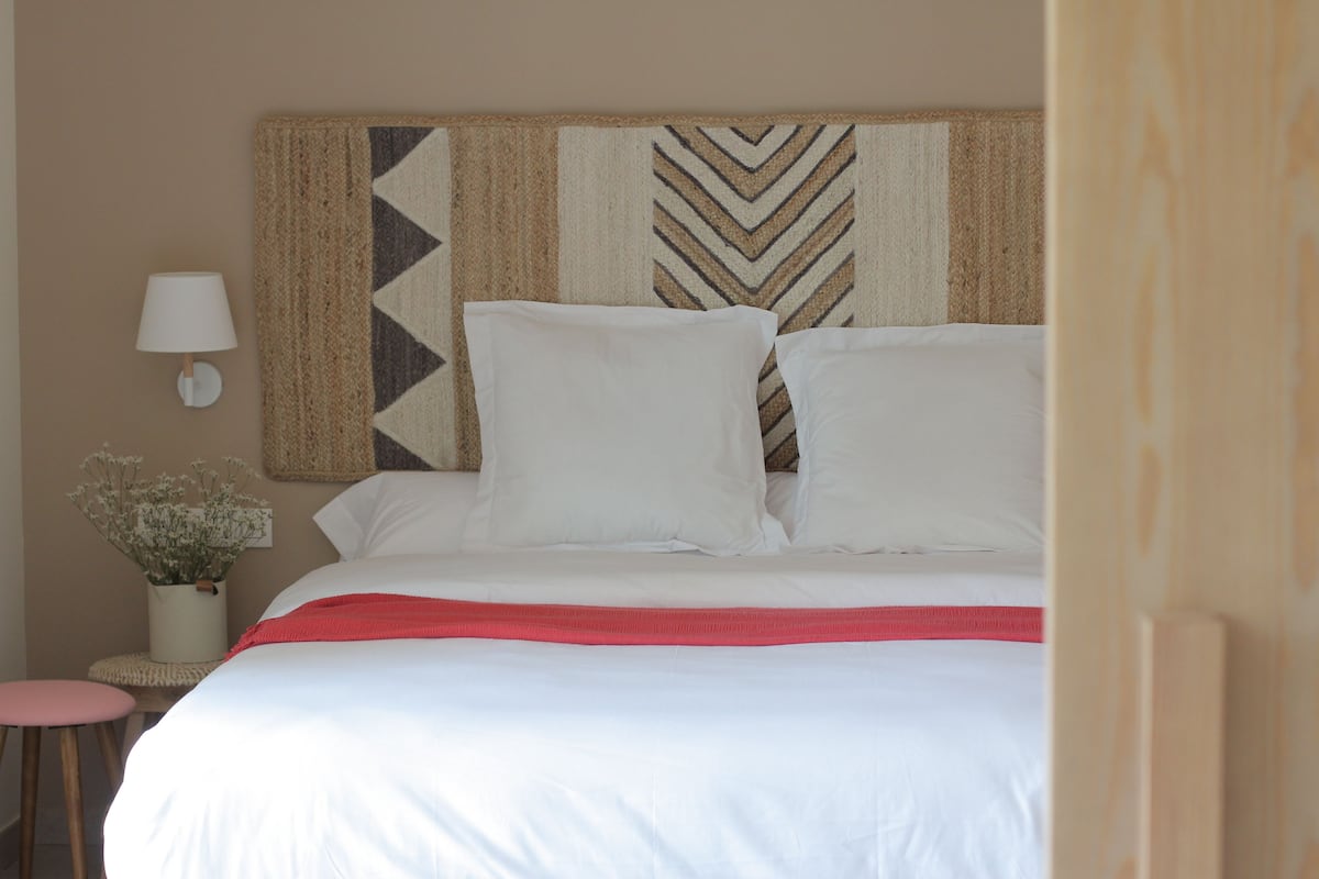A neatly made bed with white linens and a red throw blanket, a wall-mounted lamp, and a decorative woven headboard.