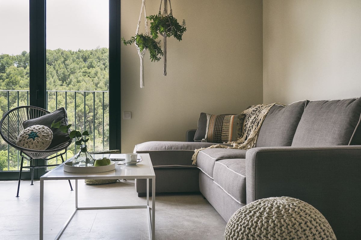 A modern living room with a grey sofa, a black wireframe chair, a small table with a glass vase and a cup, and a view of a forest through a balcony railing.