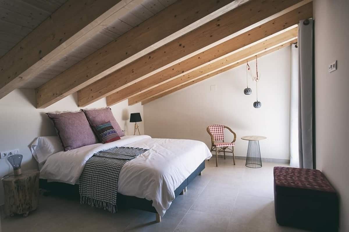 A bedroom with a sloped ceiling, a bed with white bedding, a chair, a small round table, and a bench.