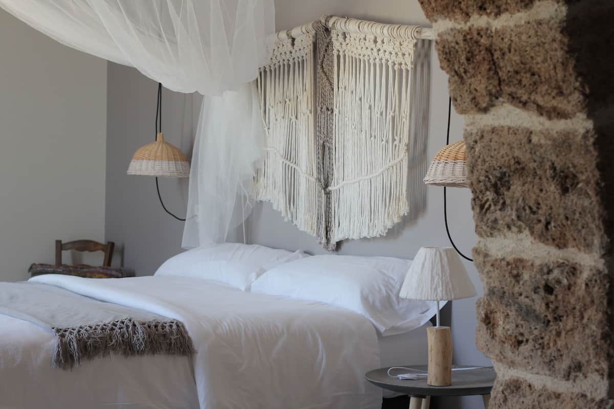 A bedroom with a bed, white bedding, a macrame headboard, a bedside lamp, and a wicker lamp.