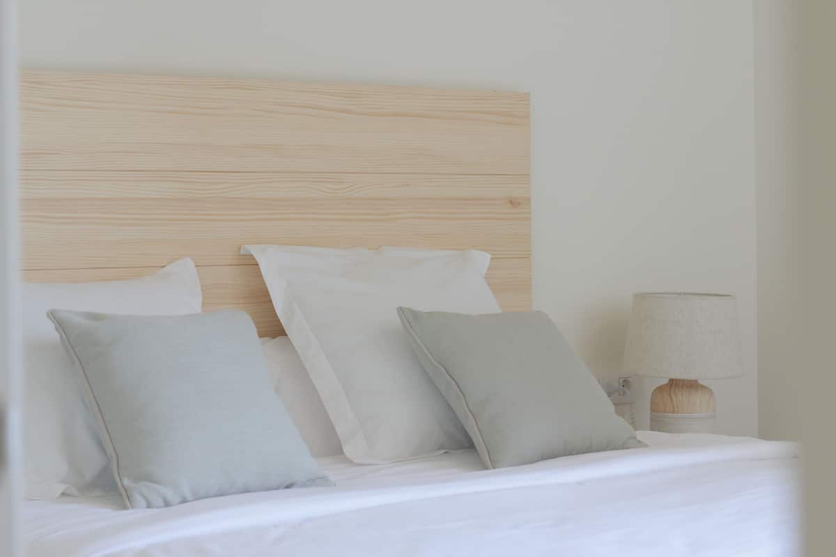 A neatly made bed with white linens and two pillows, a wooden headboard, and a bedside lamp.