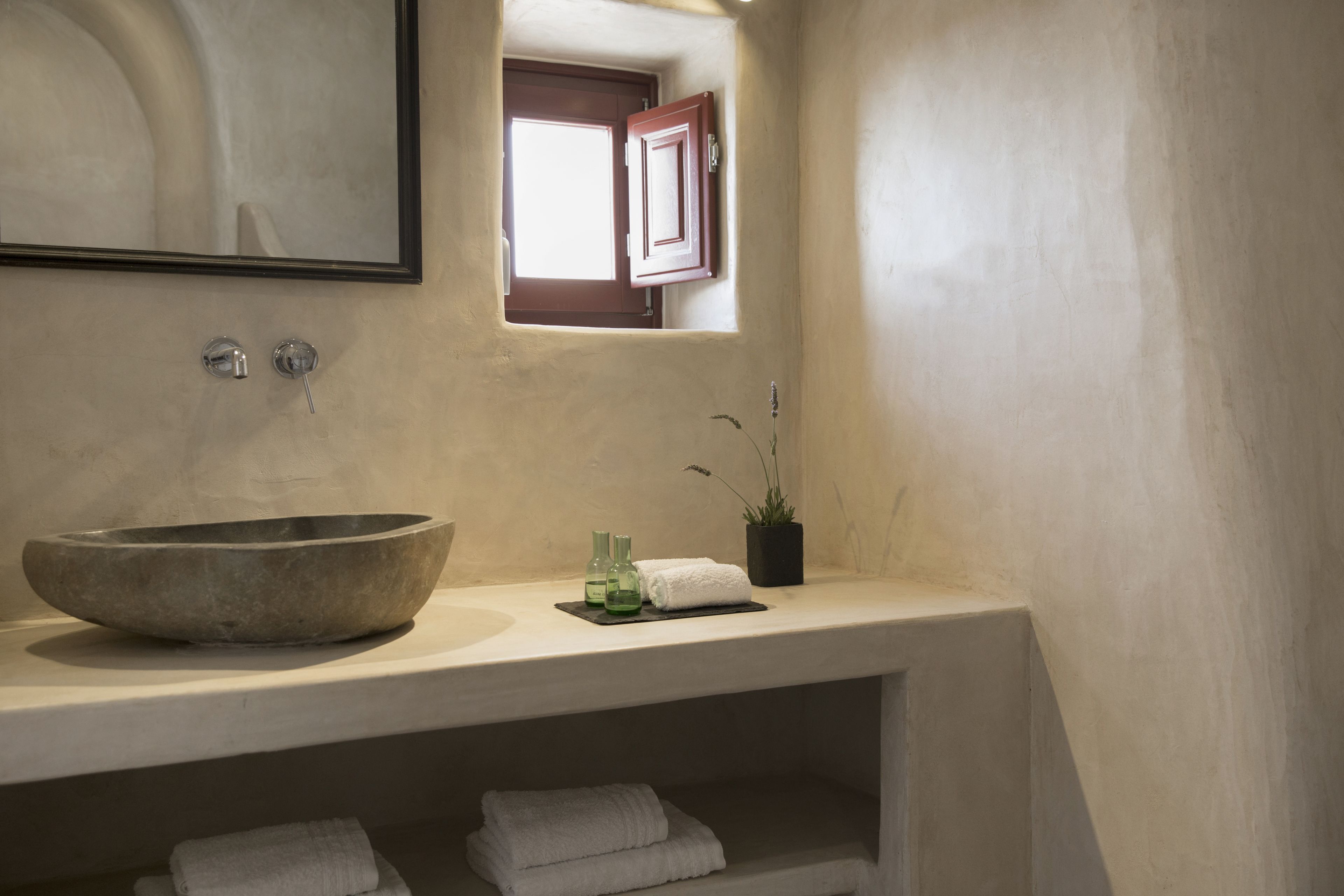 A bathroom with a large stone sink, a mirror, a window with a wooden door, and a shelf with towels and a potted plant.