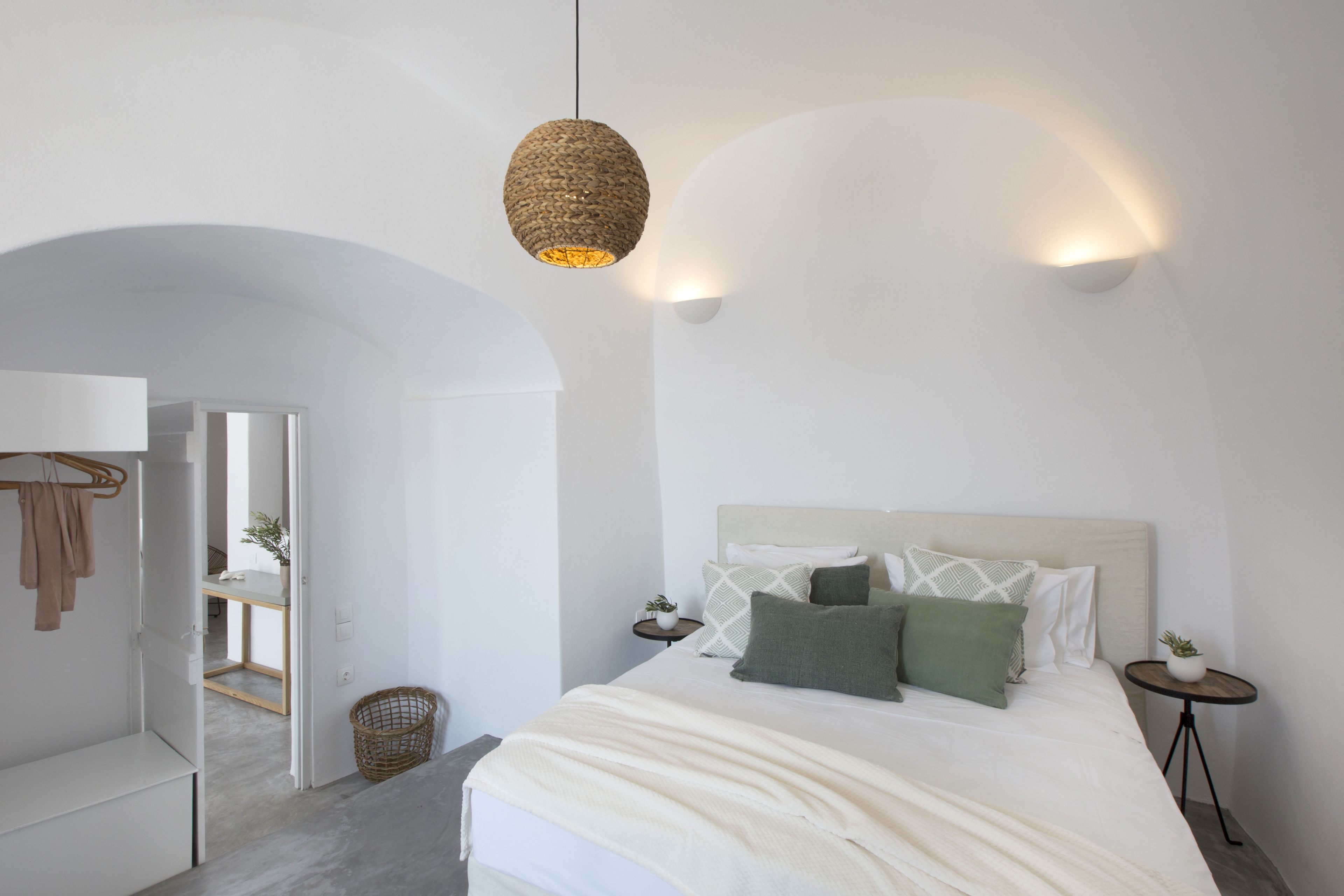 Un dormitorio minimalista con una cama grande, dos mesitas de noche, una lámpara colgante y un acceso a otra habitación.