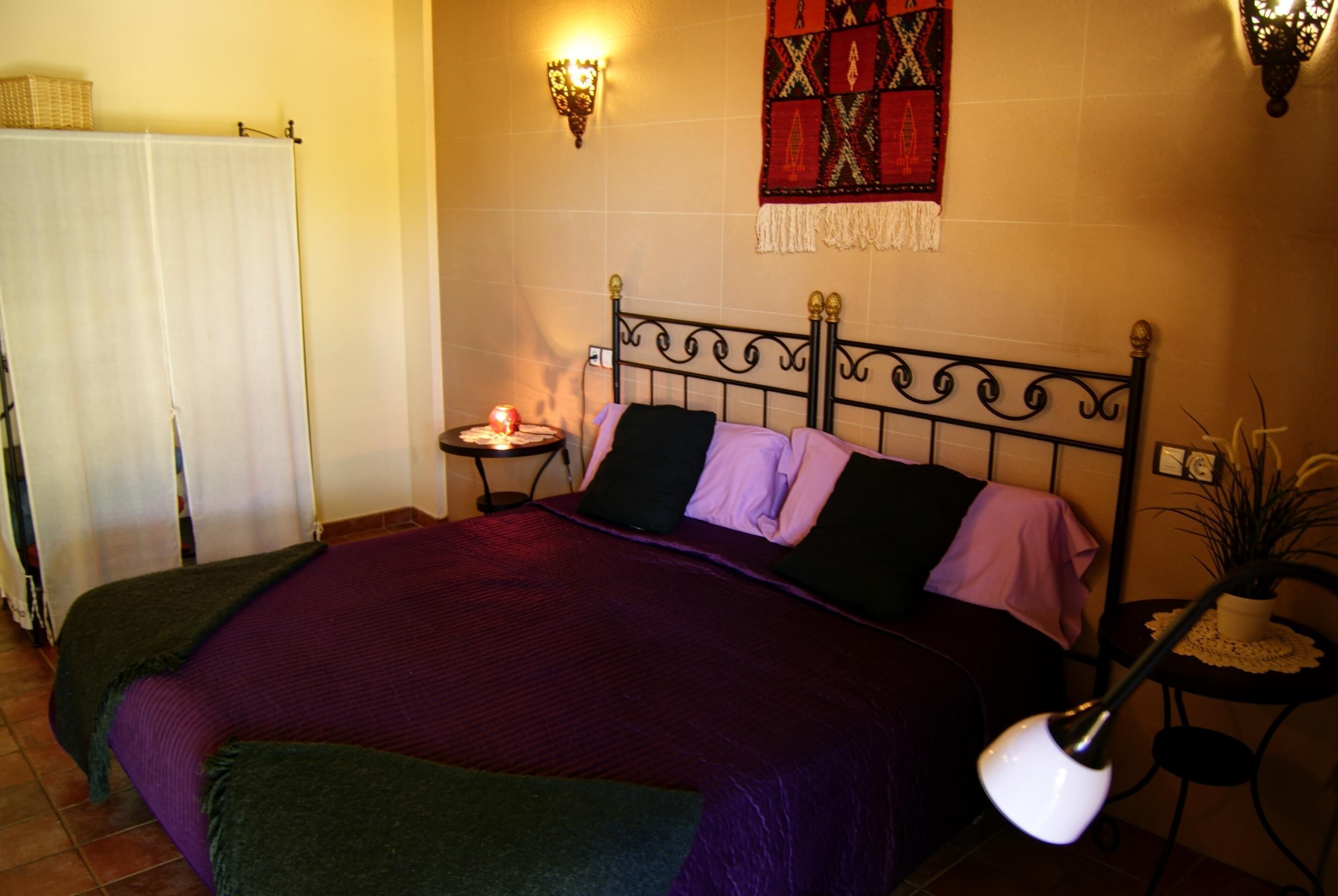 Un lit bien fait, avec une couvre-lit et des oreillers violets, une table de chevet avec une lampe et une tapisserie accrochée au mur.
