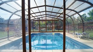 A covered swimming pool with a glass enclosure, surrounded by a patio area with lounge chairs.