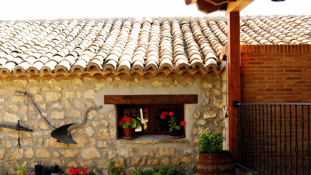 A stone building with a tiled roof, a wooden door, and a small window with red flowers. There are potted plants outside.