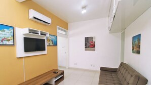Een woonkamer met een flatscreen televisie aan een witte muur, een bruine bank en een houten bankje.