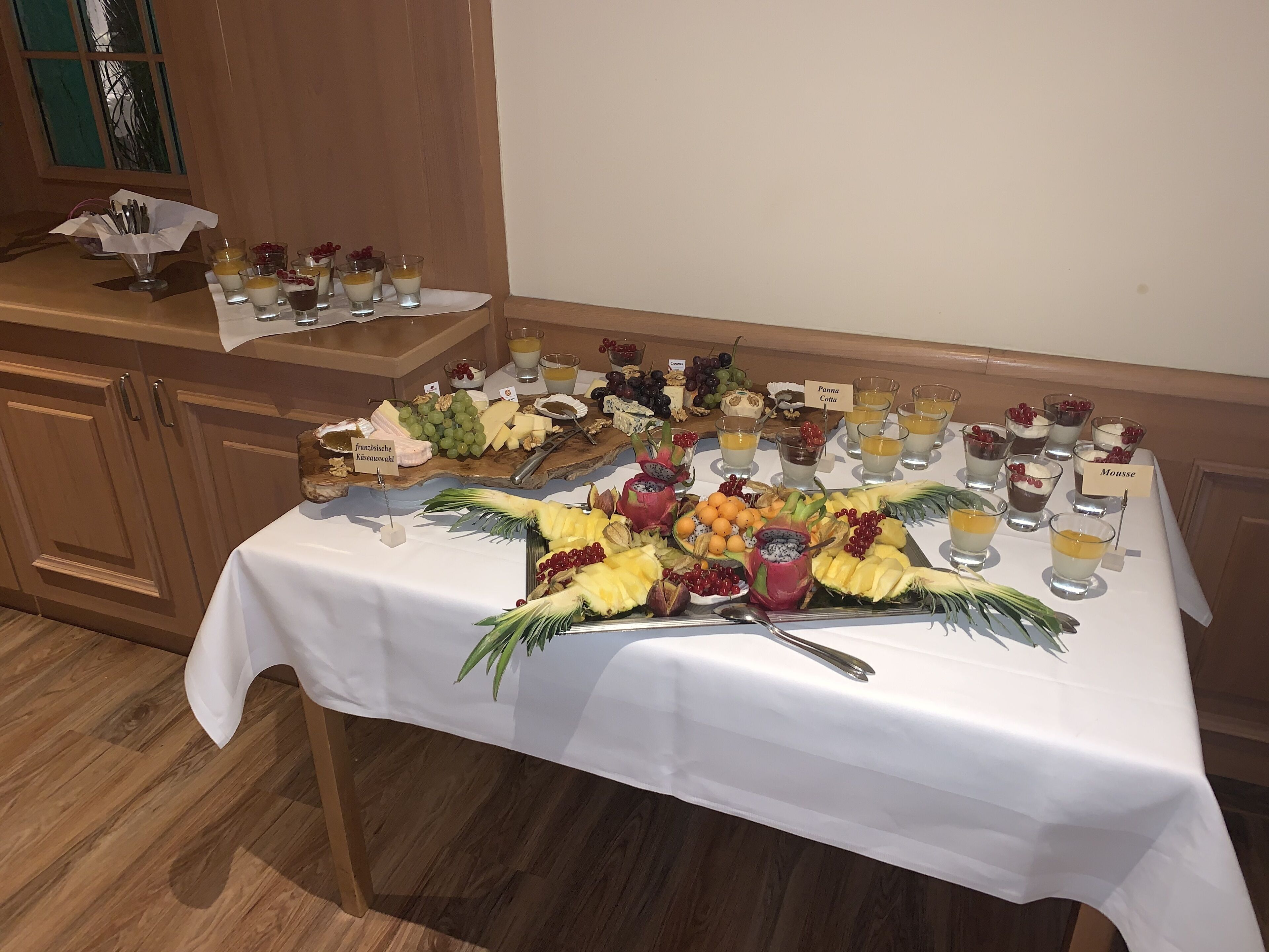 Een buffet tafel met diverse etenswaren en drankjes, waaronder fruit, kazen en desserts, uitgestald op een witte tafelkleed.