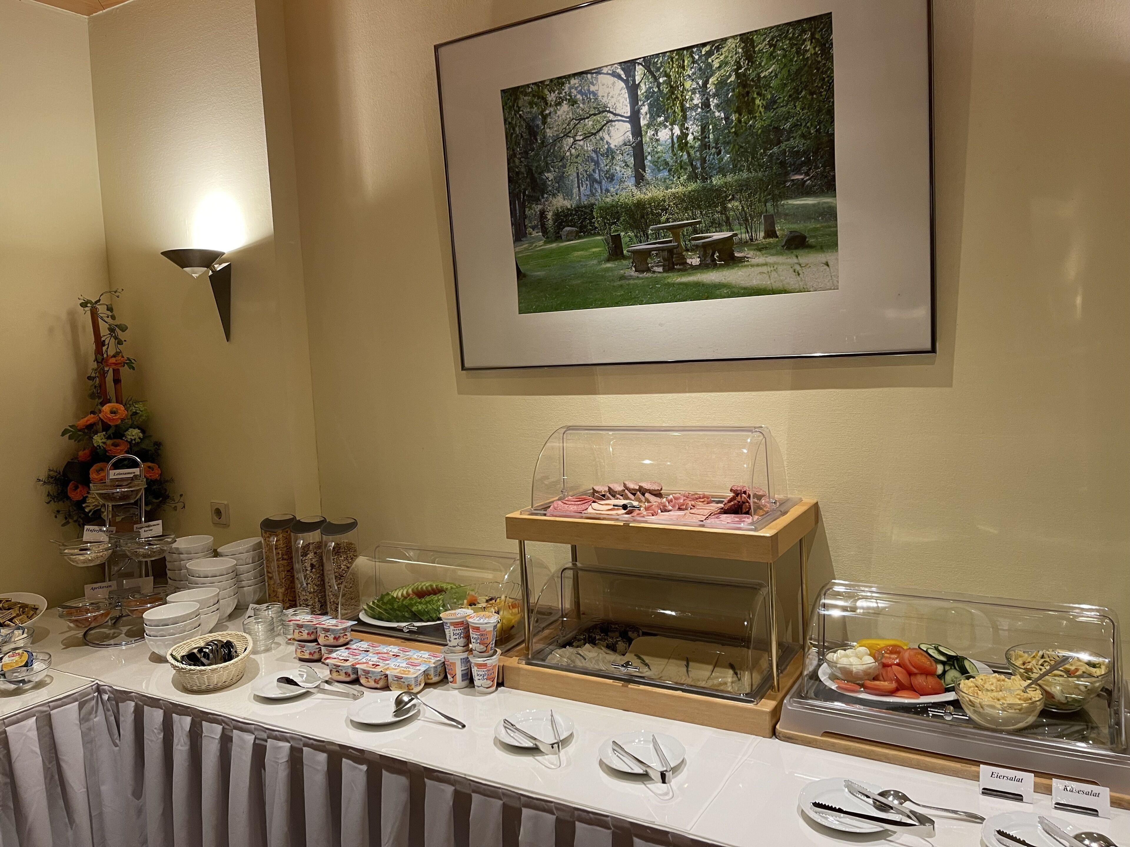 Een buffetopstelling met diverse gerechten en bestek, een lijst met een foto van een bos aan de muur en een decoratieve bloemstelling.