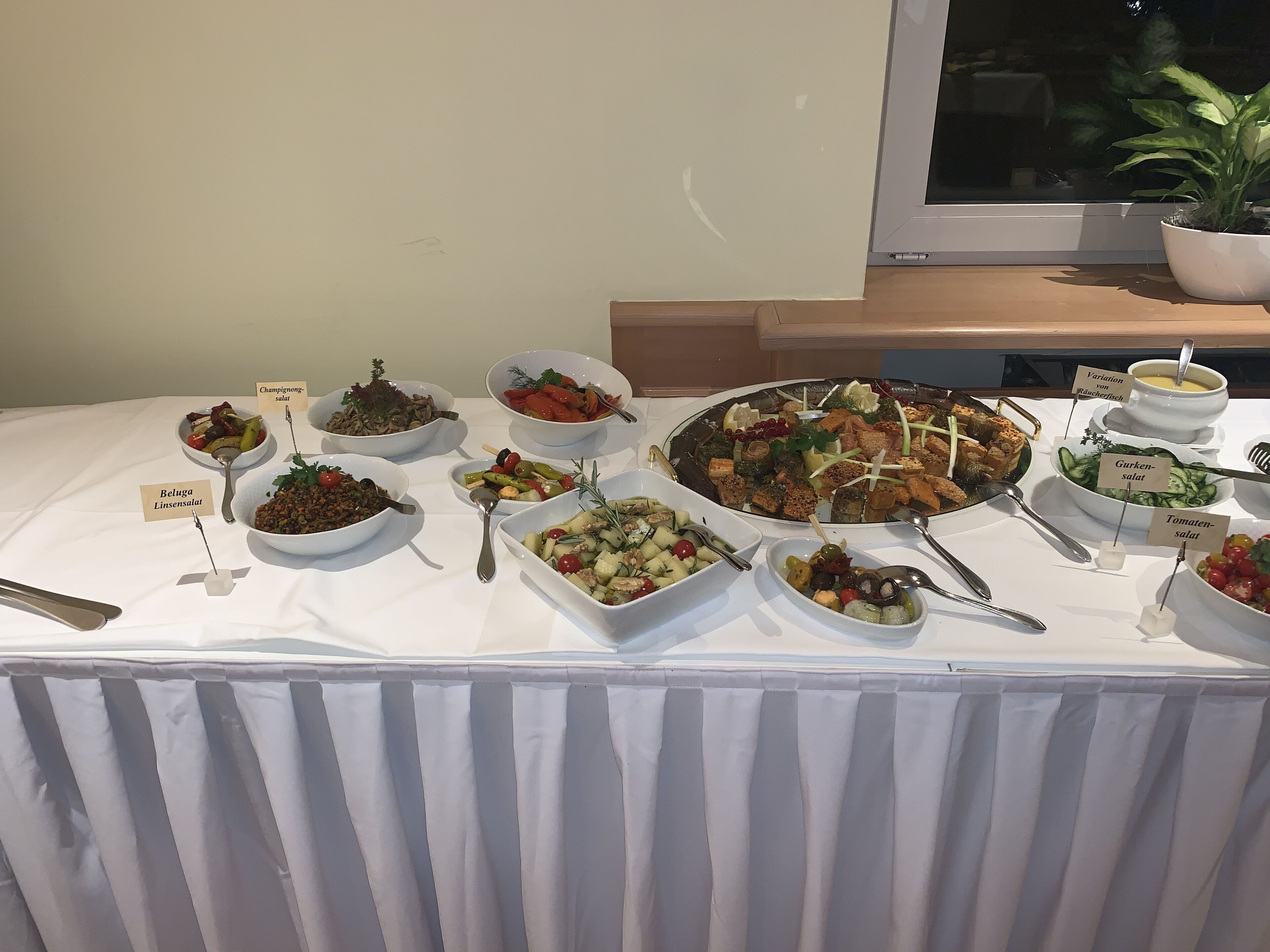 Una mesa de buffet con varios platos etiquetados en múltiples idiomas, incluyendo ensalada de lentejas Beluga, ensalada de champiñones y ensalada de pepinos.
