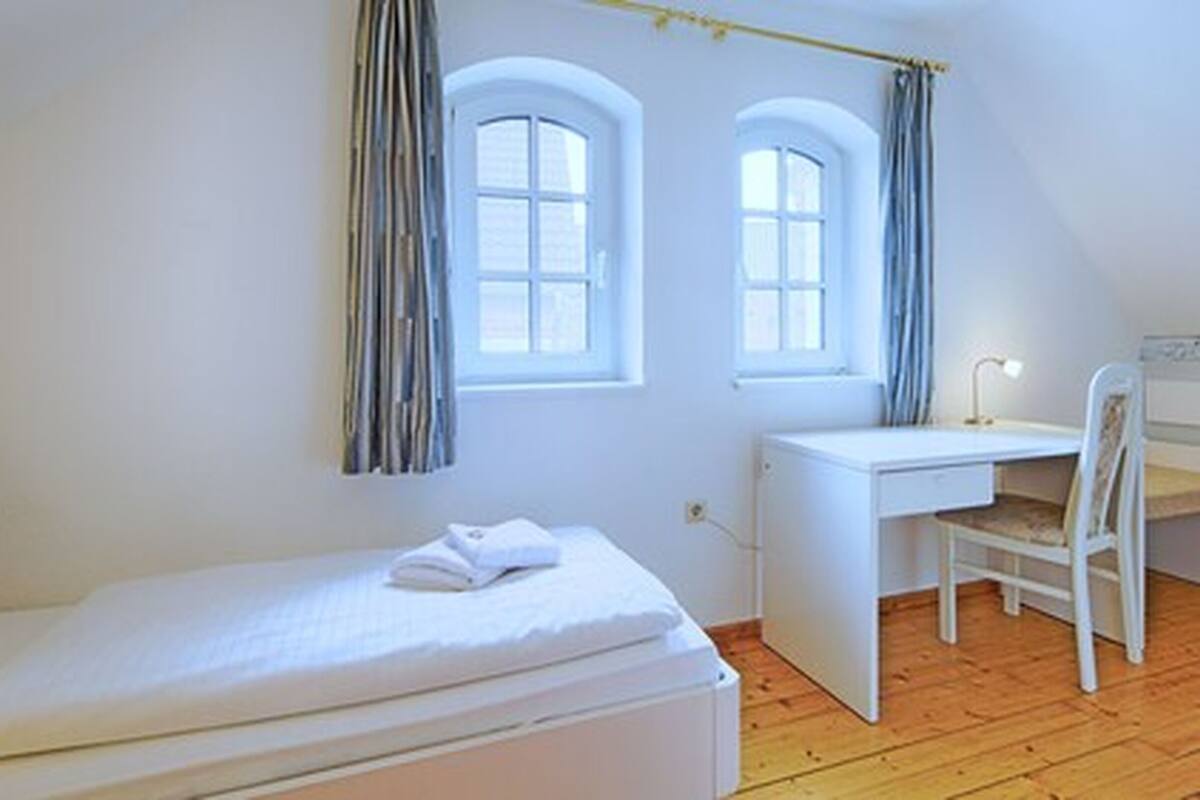 Un dormitorio con una cama individual, un escritorio y dos ventanas con cortinas.