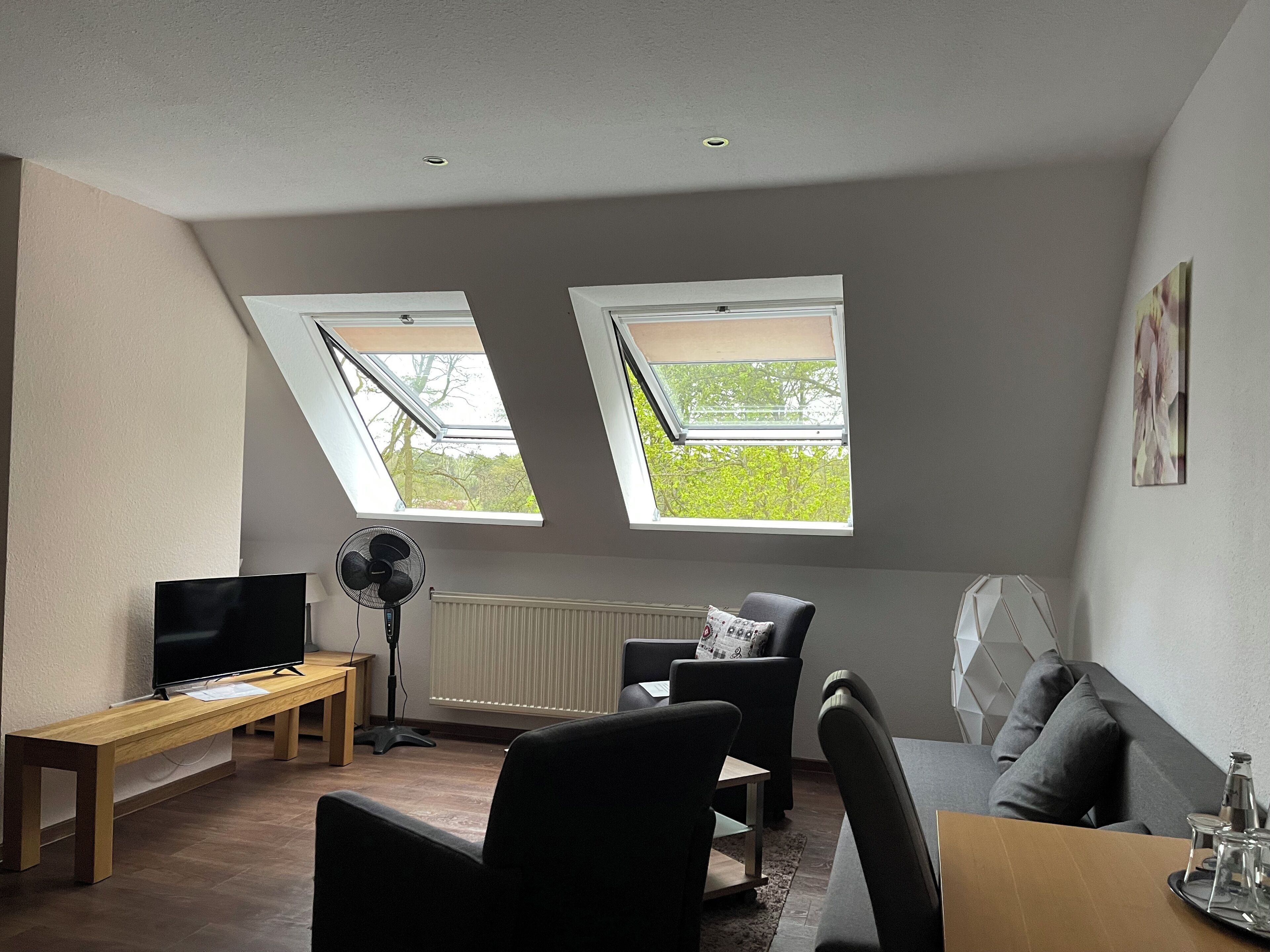 Ein modernes Wohnzimmer mit Flachbildfernseher, einer Holzbank, zwei schwarzen Sesseln, einem kleinen Tisch und einer Couch. Zwei Dachfenster sorgen für natürliches Licht und bieten Blick ins Grüne.