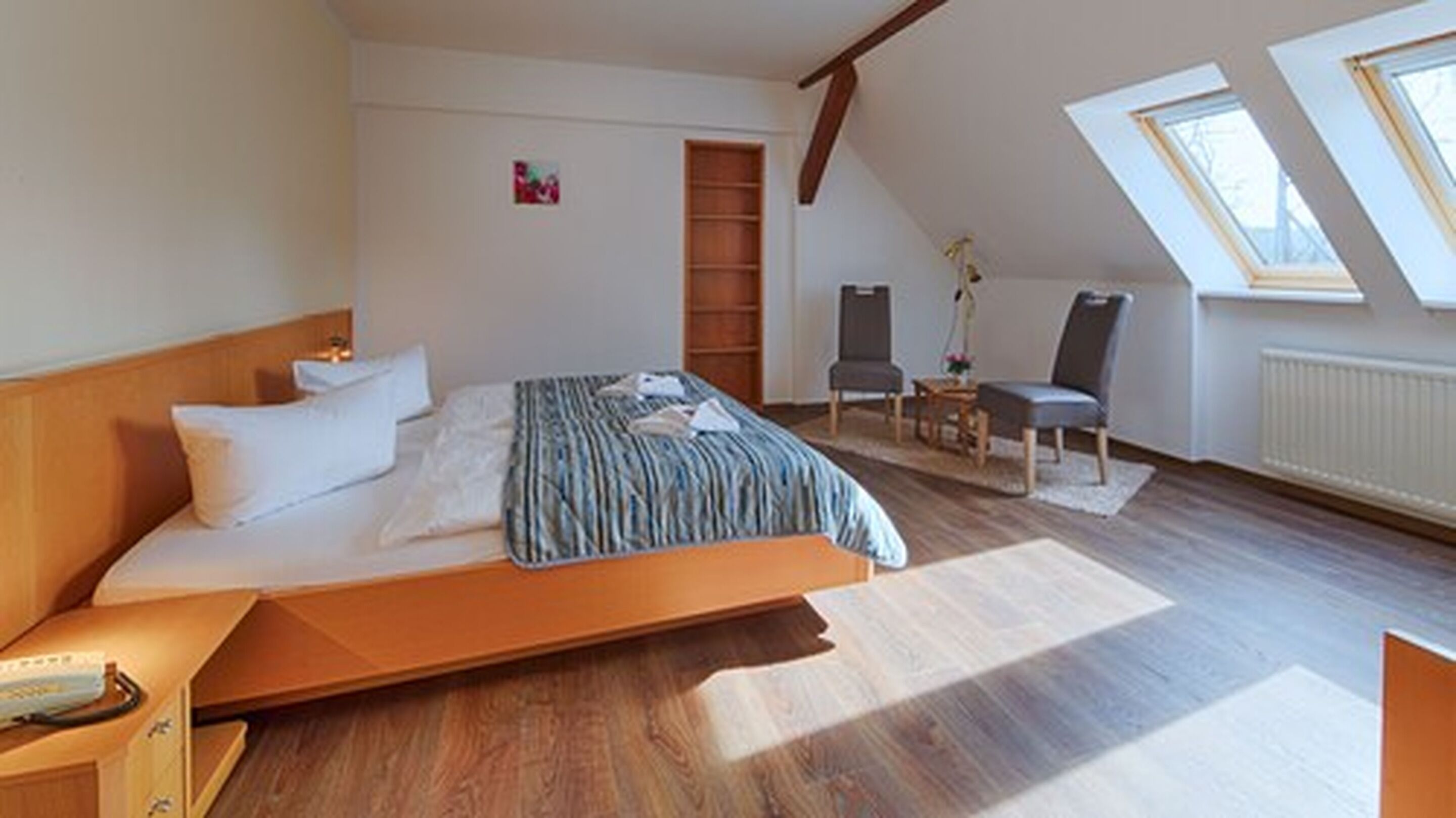Ein modernes Schlafzimmer mit einem großen Bett, einem Holz-Kopfteil, einem kleinen Schreibtisch und einem Stuhl. Es gibt ein Oberlicht und eine Heizung an der Wand.