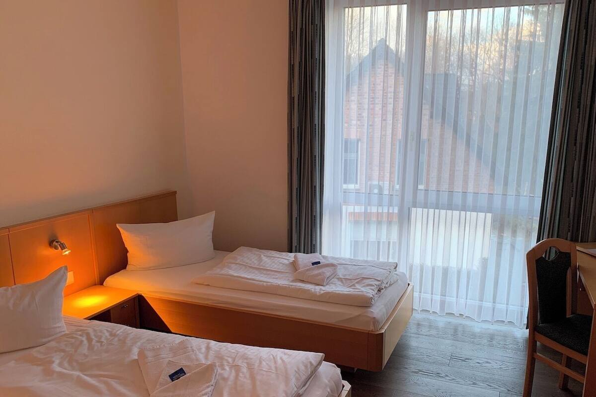 Habitación de hotel con dos camas, cabecera de madera, mesita de noche con lámpara y vistas a un edificio a través de cortinas translúcidas.