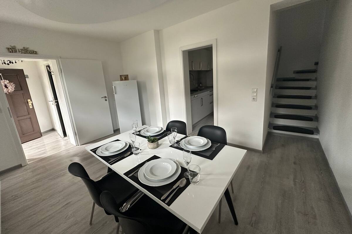 Comedor con una mesa blanca para cuatro, sillas negras y una cocina visible a través de una puerta abierta.